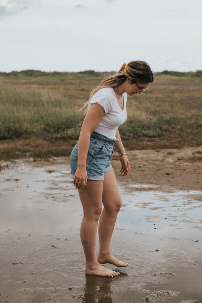 A woman in a white T-shirt and denim shorts stands barefoot on a muddy patch of ground reminiscent of Cleethorpes beach. Her hair is tied back, and she's gazing down at the earth. In the background, a grassy field stretches under a cloudy sky. © Aimee Lince Photography - Wedding photographer in Lincolnshire, Yorkshire & Nottinghamshire