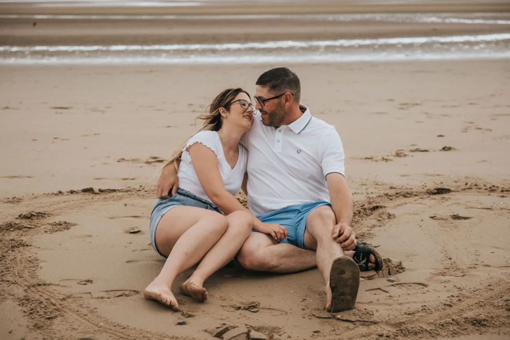 A couple sits on the sand at Cleethorpes beach, smiling at each other. The man, wearing a white polo and blue shorts, holds sandals in one hand. The woman wears a white top and denim shorts. Waves and wet sand stretch out behind them under a cloudy sky. © Aimee Lince Photography - Wedding photographer in Lincolnshire, Yorkshire & Nottinghamshire