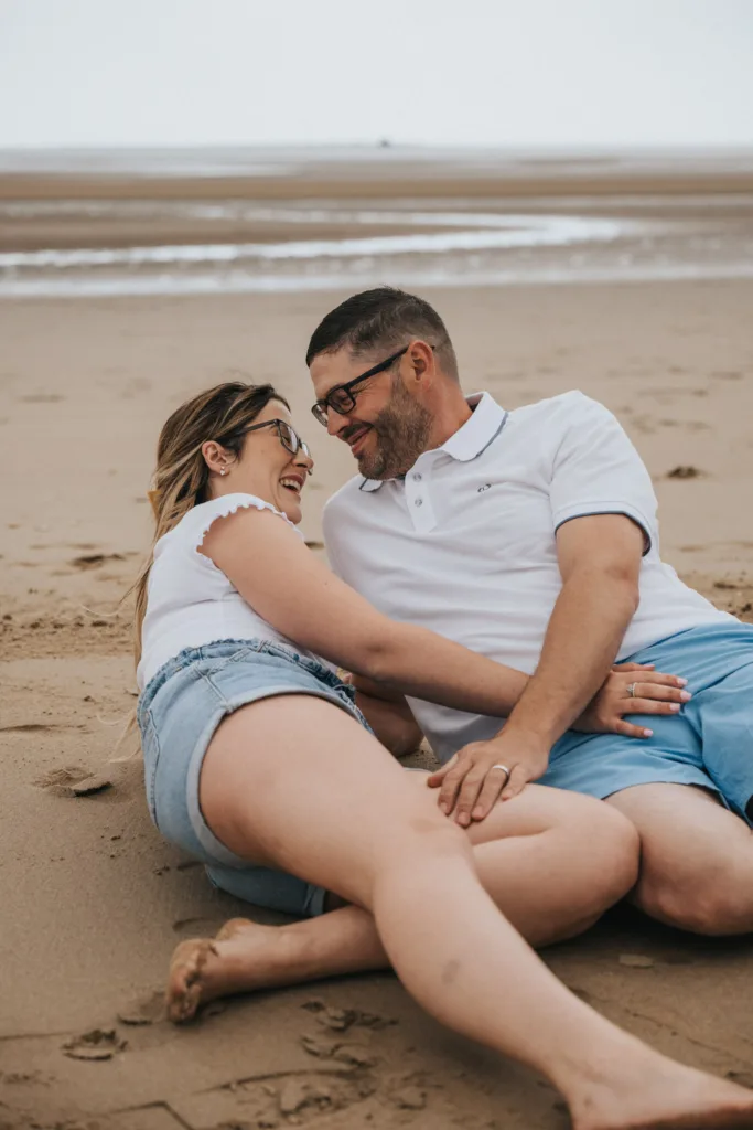 A couple relaxes on Cleethorpes Beach. The woman, wearing a white top and denim shorts, is lying close to the man. The man, in a white polo and blue shorts, reclines beside her. Both are smiling and looking at each other affectionately, with the sea visible in the background. © Aimee Lince Photography - Wedding photographer in Lincolnshire, Yorkshire & Nottinghamshire