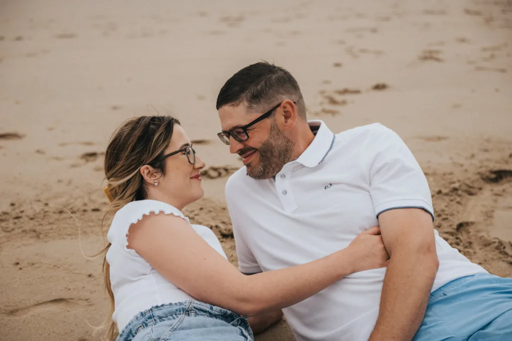 A couple, both wearing glasses, lies on the smooth, undisturbed sand of Cleethorpes Beach facing each other. The woman has long hair, a white shirt, and shorts; the man sports short hair, a beard, a white polo, and blue shorts. They smile blissfully, embodying relaxation and contentment by the shore. © Aimee Lince Photography - Wedding photographer in Lincolnshire, Yorkshire & Nottinghamshire
