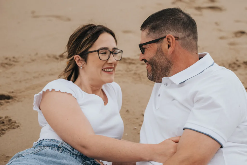 A couple relaxes on Cleethorpes beach, smiling at each other beneath their glasses. Both sport white tops—the woman in a short-sleeve shirt and denim shorts, the man in a crisp polo with long pants. Behind them, smooth sand captures a few wandering footprints. © Aimee Lince Photography - Wedding photographer in Lincolnshire, Yorkshire & Nottinghamshire