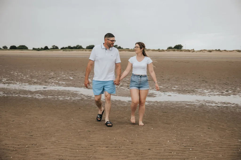 A couple walks hand in hand along Cleethorpes beach. The man wears a white polo shirt, blue shorts, and sandals, while the woman sports a white t-shirt and denim shorts, barefoot. The sky is overcast, with a line of trees visible in the distance. They appear to be smiling and talking. © Aimee Lince Photography - Wedding photographer in Lincolnshire, Yorkshire & Nottinghamshire