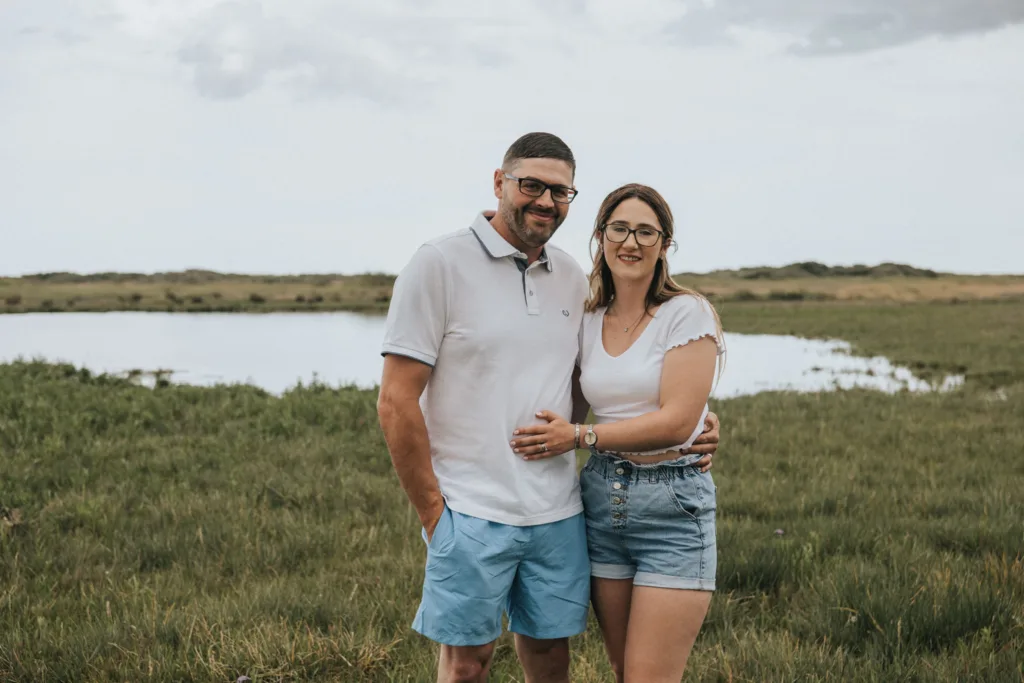 A couple stands on green grass near a calm body of water under a cloudy sky at Cleethorpes beach. The man wears glasses, a white polo shirt, and blue shorts with one hand in his pocket. The woman also sports glasses, a white shirt, and denim shorts with her arm around him. They both smile at the camera. © Aimee Lince Photography - Wedding photographer in Lincolnshire, Yorkshire & Nottinghamshire
