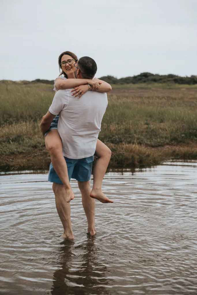 A man carries a woman in his arms while standing in the shallow waters of Cleethorpes beach. The woman, smiling and wearing glasses, sports a white top and denim shorts. The man is clad in a white shirt and blue shorts. They are surrounded by grass and open landscape under a cloudy sky. © Aimee Lince Photography - Wedding photographer in Lincolnshire, Yorkshire & Nottinghamshire