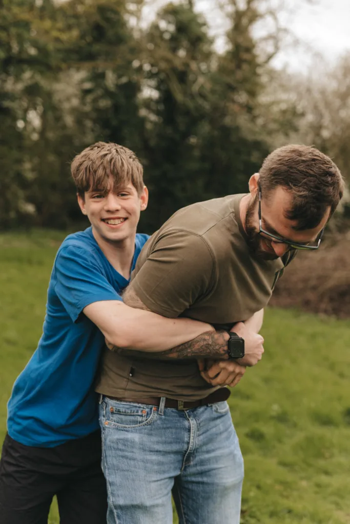Two people are playfully embracing outdoors at Hall Farm Hotel. The person on the left, wearing a blue shirt, smiles at the camera while hugging the other person from behind. The person on the right, in a green shirt and glasses, is slightly bent forward against lush greenery. © Aimee Lince Photography - Wedding photographer in Lincolnshire, Yorkshire & Nottinghamshire