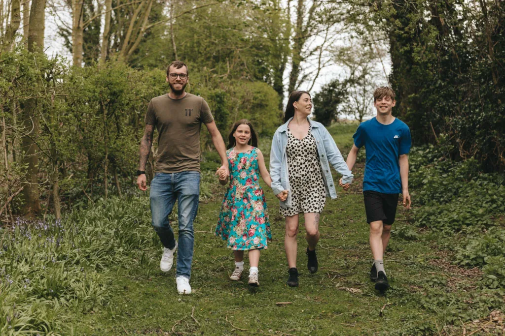 A family of four strolls hand in hand along a lush, tree-lined path near Hall Farm Hotel. The man wears a green t-shirt and jeans, the woman a denim jacket and dress, the girl a floral dress, and the teenage boy sports a blue t-shirt and black shorts, all enjoying their day together. © Aimee Lince Photography - Wedding photographer in Lincolnshire, Yorkshire & Nottinghamshire