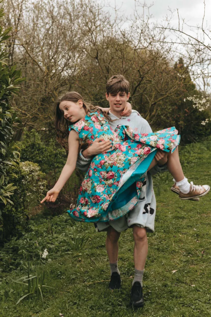 A young boy joyfully carries a girl in a vibrant floral dress through the grassy garden of Hall Farm Hotel. The girl, with long hair, looks playfully to the side. The boy, in a gray hoodie and shorts, beams with happiness. Surrounded by trees and bushes, they radiate family warmth and carefree joy. © Aimee Lince Photography - Wedding photographer in Lincolnshire, Yorkshire & Nottinghamshire