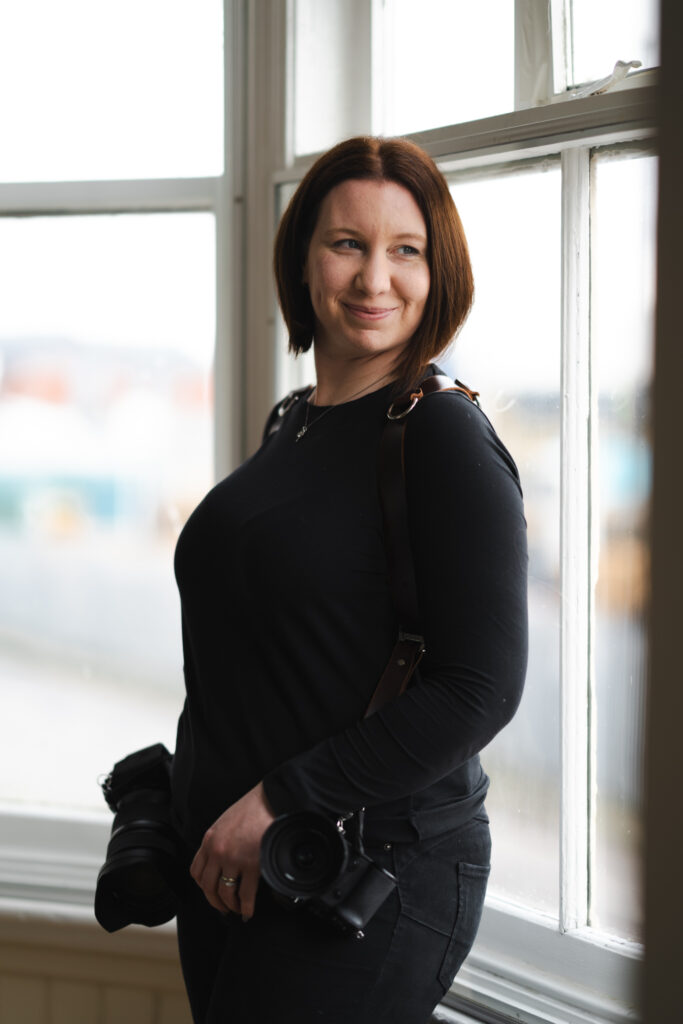 A person with shoulder-length brown hair stands by a sunlit window, smiling slightly to the side. They are dressed in a black long-sleeve shirt and black pants, wearing dual camera harnesses with cameras attached—a perfect example of how to choose a wedding photographer. The background is softly blurred, featuring light colors and soft focus. © Aimee Lince Photography