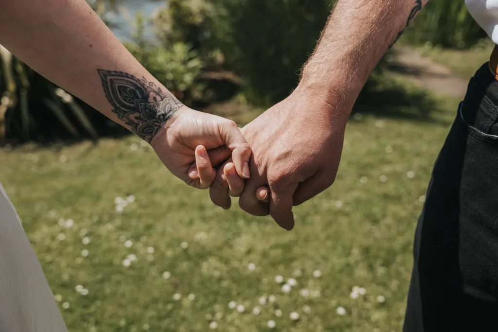 Close-up of two people holding hands outdoors on grass, capturing an intimate moment perfect for engagement photography. The left hand showcases a detailed floral tattoo, while the right is unadorned. Both wear casual clothing, surrounded by greenery and a sunlit path. © Aimee Lince Photography - Wedding photographer in Lincolnshire, Yorkshire & Nottinghamshire