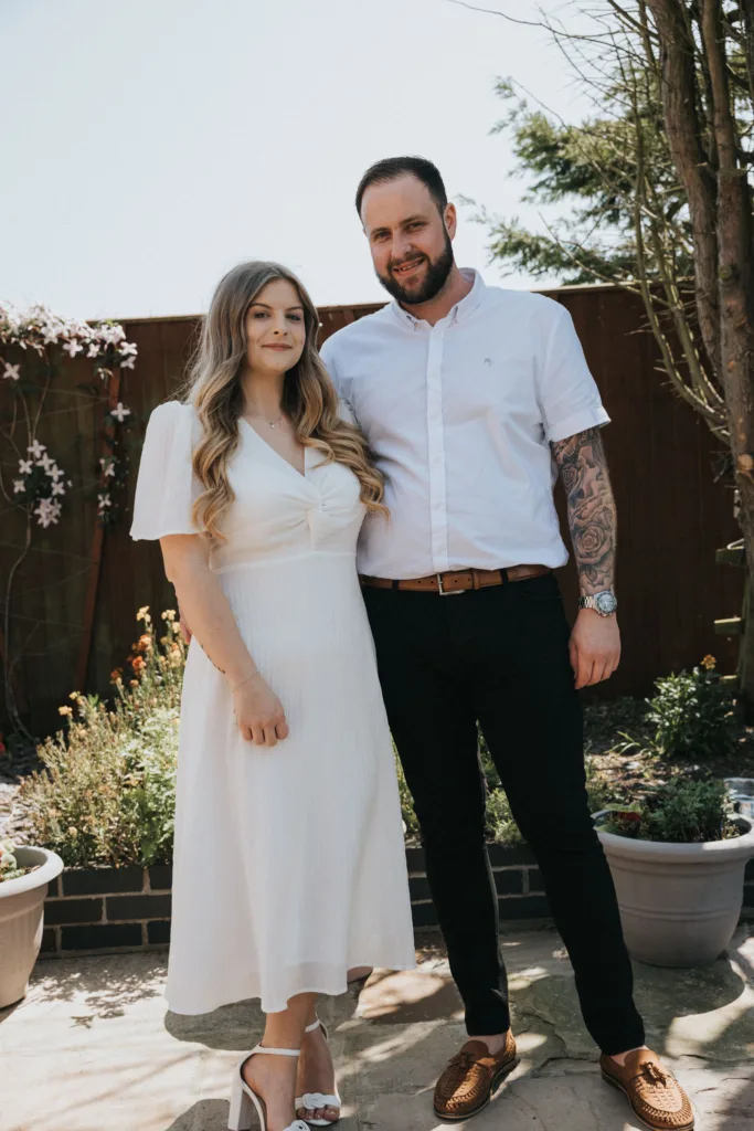 A couple poses for engagement photography in a garden. The woman wears a white dress and heels, her long, wavy hair cascading down. The man sports a white shirt and black pants, showcasing his tattooed arm. They stand on a stone path, framed by potted plants and a wooden fence. © Aimee Lince Photography - Wedding photographer in Lincolnshire, Yorkshire & Nottinghamshire