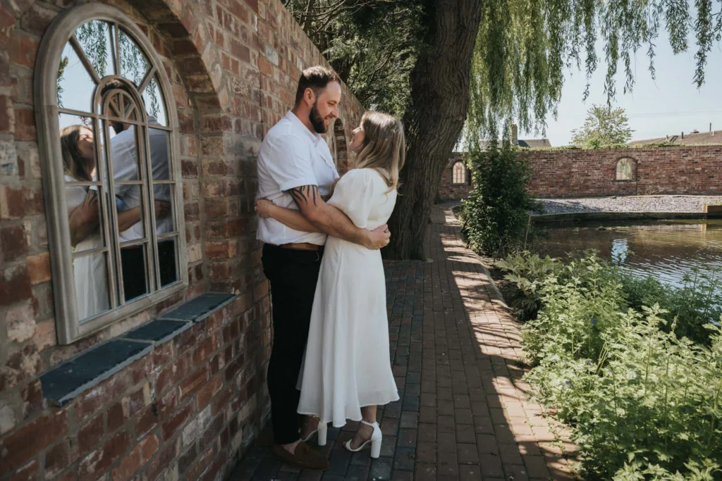 Lincolnshire Engagement Photography - Natasha & Dale © Aimee Lince Photography 21-05-23 55 of 1111 A couple embraces on a brick pathway beside a pond, perfect for engagement photography. The man in a white shirt and brown pants, and the woman in a flowing white dress share a moment as sunlight filters through the trees, casting soft shadows near the arched window of the brick wall. © Aimee Lince Photography - Wedding photographer in Lincolnshire, Yorkshire & Nottinghamshire