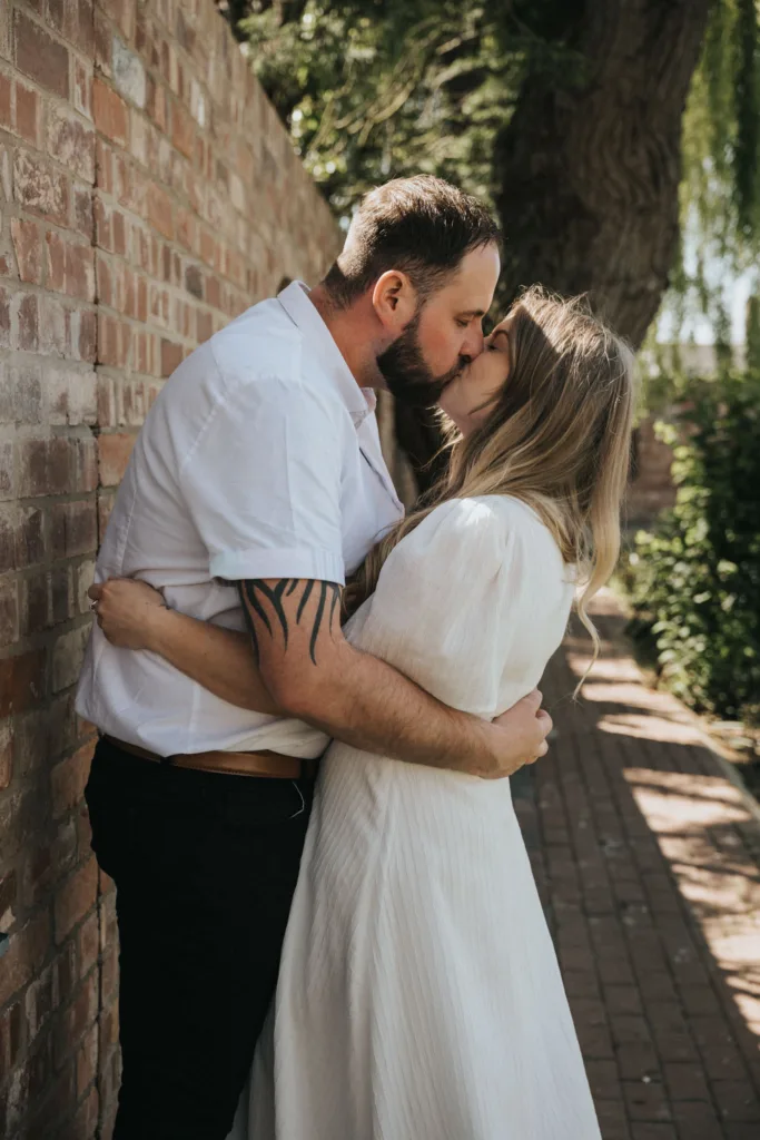 A couple shares a passionate kiss against a brick wall on a sunlit path, perfect for engagement photography. The man in a white shirt and dark pants embraces the woman in a white dress. Sunlight filters through trees, casting dappled shadows on the path and wall, creating a romantic and serene scene. © Aimee Lince Photography - Wedding photographer in Lincolnshire, Yorkshire & Nottinghamshire