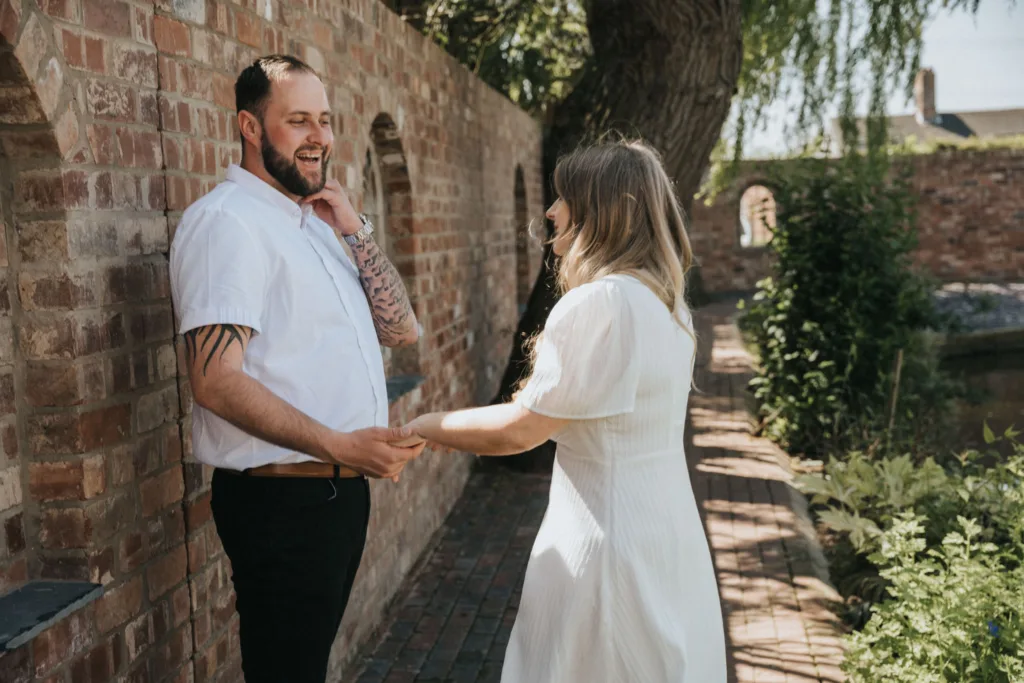 A couple stands on a brick path by a rustic wall under a large tree, captured in an enchanting moment of engagement photography. The tattooed man with a beard wears a white shirt and black pants, while the woman in a white dress has long hair. They hold hands and smile, framed by lush green foliage. © Aimee Lince Photography - Wedding photographer in Lincolnshire, Yorkshire & Nottinghamshire