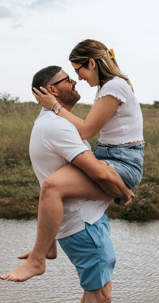 A man in sunglasses, a white shirt, and blue shorts lifts a woman in a white top and denim shorts by the water during their engagement photoshoot. She wears sunglasses and a large hair clip. They smile at each other, surrounded by grass and a cloudy sky. © Aimee Lince Photography - Wedding photographer in Lincolnshire, Yorkshire & Nottinghamshire