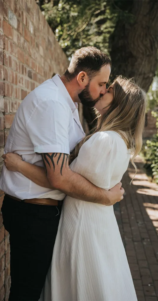 A man and woman share a kiss in a sunlit outdoor setting, perfect for engagement photography. The man wears a short-sleeved white shirt, showcasing a tattooed arm, while the woman is dressed in a white dress. They stand near a brick wall with lush greenery behind them, their embrace affectionate and intimate. © Aimee Lince Photography - Wedding photographer in Lincolnshire, Yorkshire & Nottinghamshire