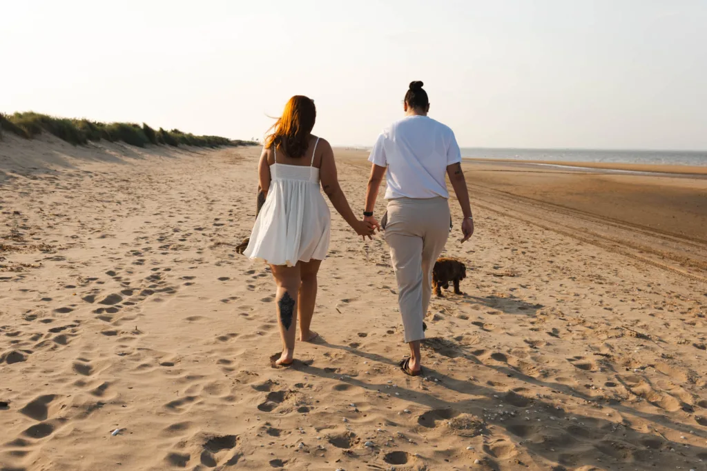 Two people wander hand in hand along Cleethorpes Beach, heading towards the horizon. One wears a flowing white dress, the other a crisp white shirt and pants. A small dog trots beside them as the sun casts soft shadows on the sandy expanse, while grassy dunes line the left side of this idyllic scene. © Aimee Lince Photography - Wedding photographer in Lincolnshire, Yorkshire & Nottinghamshire