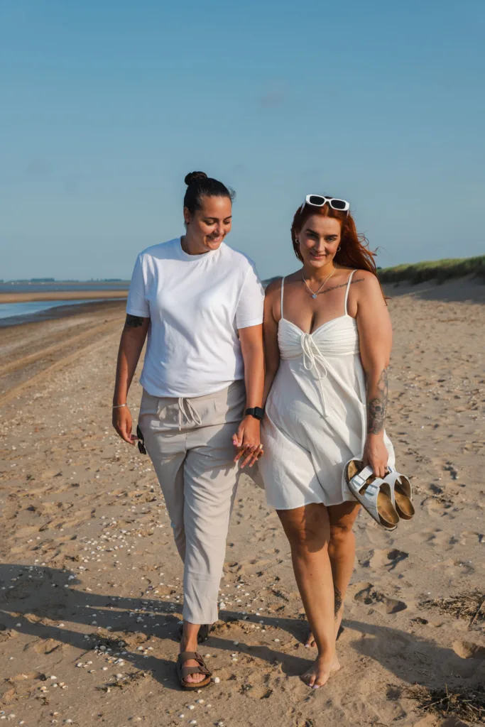 Two people walk hand in hand along Cleethorpes Beach. One wears a white t-shirt and beige pants, while the other wears a white dress and holds sandals. They are smiling under a clear blue sky. The beach is lined with shells and grasses, with the ocean in the background. © Aimee Lince Photography - Wedding photographer in Lincolnshire, Yorkshire & Nottinghamshire