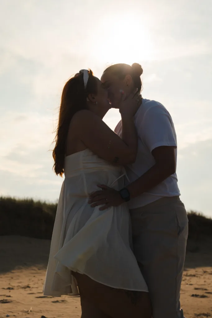 A couple is standing on Cleethorpes Beach during sunset, silhouetted by the sun. They are embracing and sharing a kiss. One wears a white dress and sunglasses on their head, the other a white shirt and beige pants. The sky is partly cloudy, and the beach is serene and dimly lit. © Aimee Lince Photography - Wedding photographer in Lincolnshire, Yorkshire & Nottinghamshire