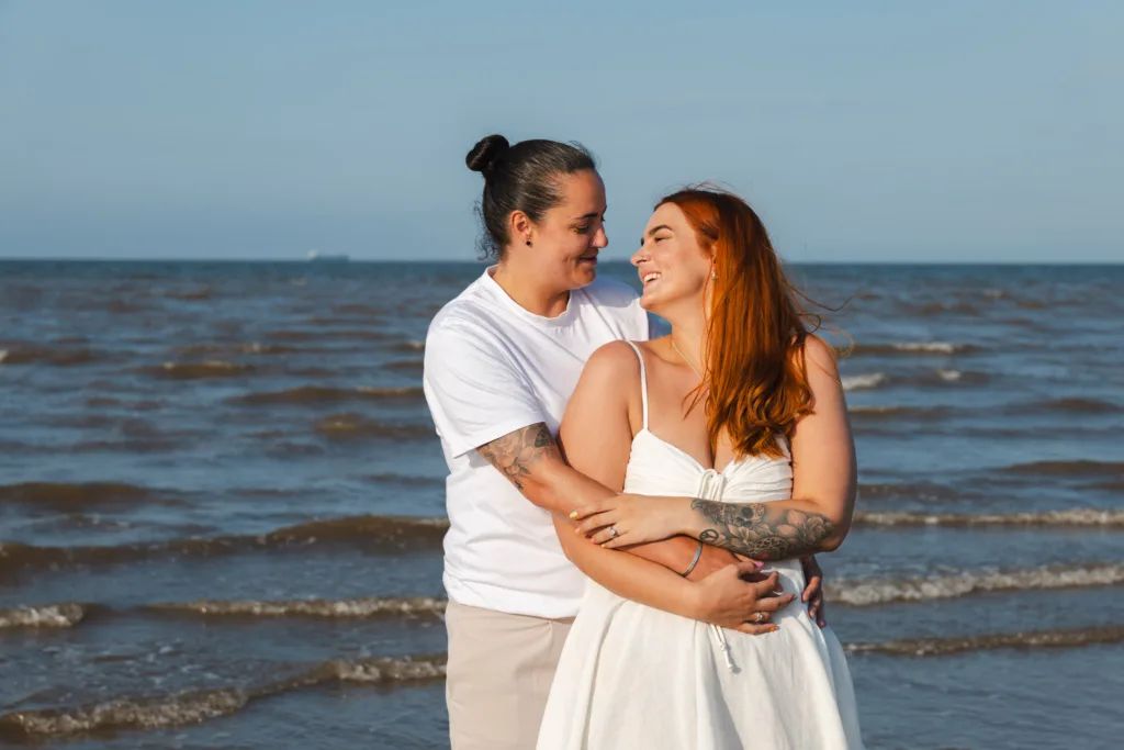 A smiling couple stands on Cleethorpes Beach, embracing. One with long red hair wears a white dress, while the other with dark hair in a bun sports a white shirt and beige shorts. Both have visible tattoos, as the ocean waves gently lap behind them under a clear blue sky. © Aimee Lince Photography - Wedding photographer in Lincolnshire, Yorkshire & Nottinghamshire