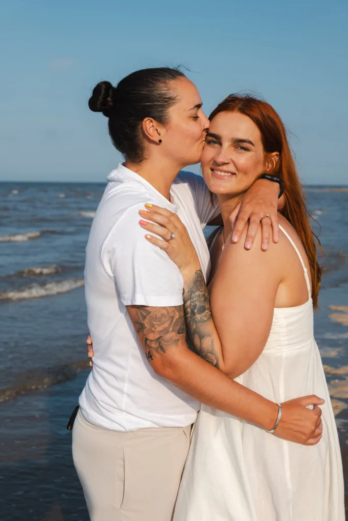 A couple embraces on Cleethorpes Beach. One person with tied-back hair and a tattooed arm kisses the other's forehead. The other smiles, wearing a white dress. With the ocean and blue sky in the background, they appear content and relaxed, enjoying a sunny day by the sea. © Aimee Lince Photography - Wedding photographer in Lincolnshire, Yorkshire & Nottinghamshire
