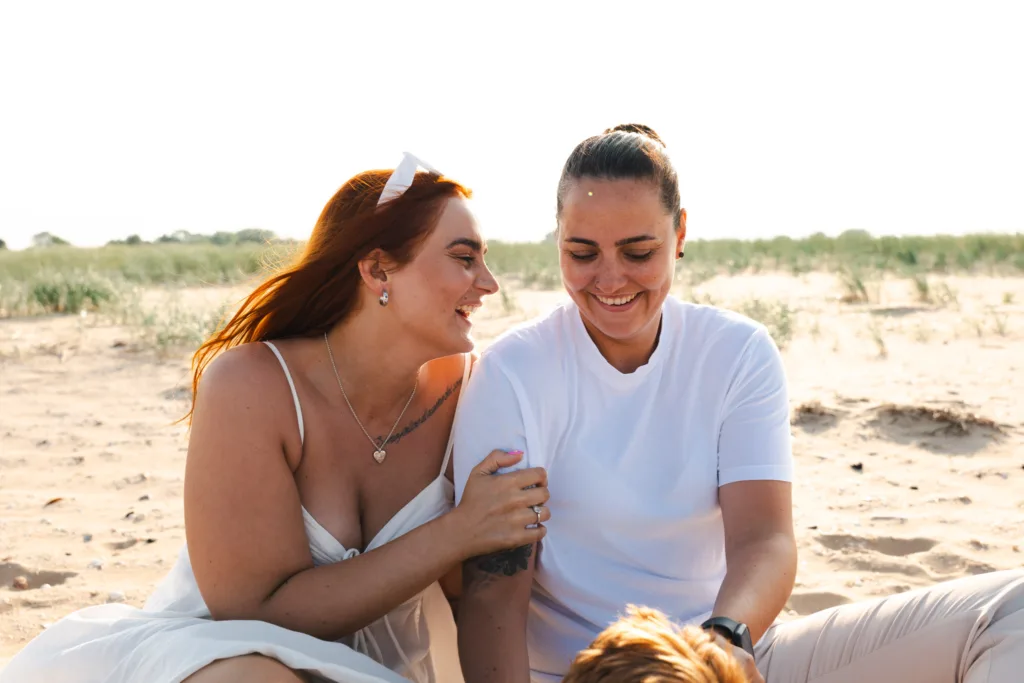 Two people sitting on Cleethorpes Beach, smiling at each other. The person on the left has long red hair and wears a white dress. The person on the right has short dark hair and wears a white t-shirt. Tall grasses are in the background under a bright sky. © Aimee Lince Photography - Wedding photographer in Lincolnshire, Yorkshire & Nottinghamshire