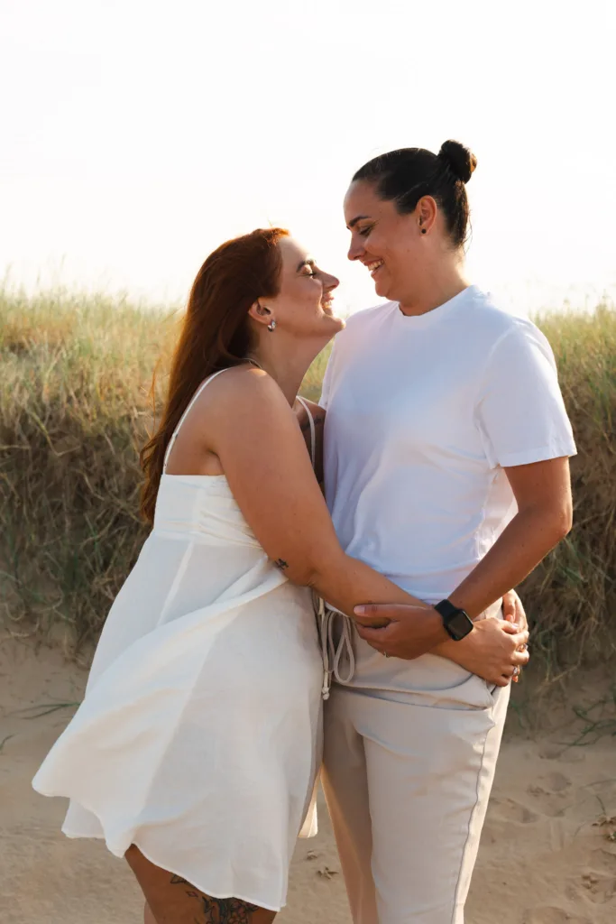 Two people stand on Cleethorpes Beach with grass swaying in the background. One with long red hair in a white dress leans towards the other, who has dark hair in a bun and wears a white t-shirt and beige pants. They smile and gaze at each other, creating a warm, intimate moment. © Aimee Lince Photography - Wedding photographer in Lincolnshire, Yorkshire & Nottinghamshire