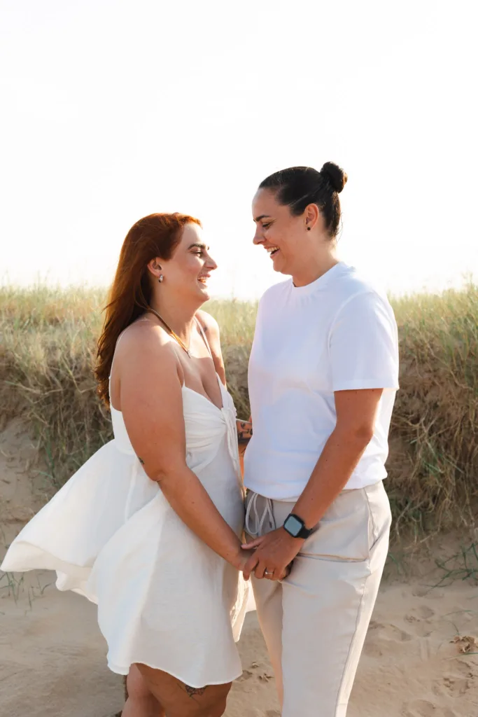Two people stand on Cleethorpes Beach with grassy dunes. The person on the left wears a white dress and long reddish hair. On the right, a bun complements their white shirt and light pants. They hold hands, smiling warmly at each other against the bright sky. © Aimee Lince Photography - Wedding photographer in Lincolnshire, Yorkshire & Nottinghamshire