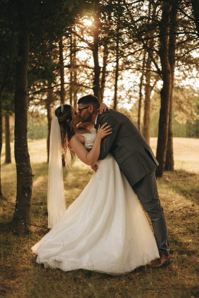 A bride and groom share a romantic kiss in a sunlit forest at Messingham Grange. The bride wears a white gown and veil, while the groom is in a gray suit. Surrounded by tall trees, the sun casts warm light through the leaves, creating a serene and intimate atmosphere. © Aimee Lince Photography