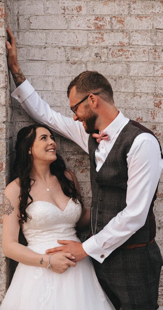 A couple stands against a brick wall in wedding attire at Hall Farm Hotel. The bride, in a strapless white gown with lace details and a necklace, smiles at the groom, who wears glasses, a gray vest, white shirt, and bow tie. They hold hands lovingly as his other arm rests against the wall. © Aimee Lince Photography