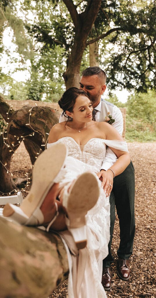 A bride and groom sit on a tree branch in the tranquil woodland setting of Hirst Priory. The bride, in an off-shoulder lace wedding dress, leans back against the groom, dressed casually in a light shirt and dark pants. Her white heels are propped up as twinkling lights decorate the tree behind them. © Aimee Lince Photography