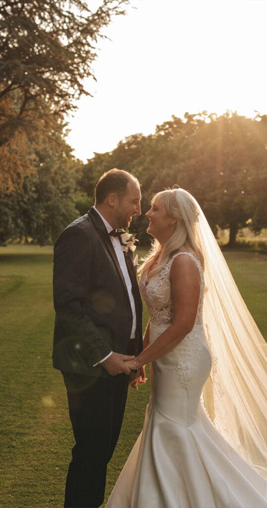 A bride in a white gown and veil, and a groom in a black suit and bow tie, stand hand in hand on the grassy grounds of Wootton Hall. They gaze into each other's eyes, illuminated by the warm glow of the setting sun behind them, with trees gracefully framing their moment. © Aimee Lince Photography