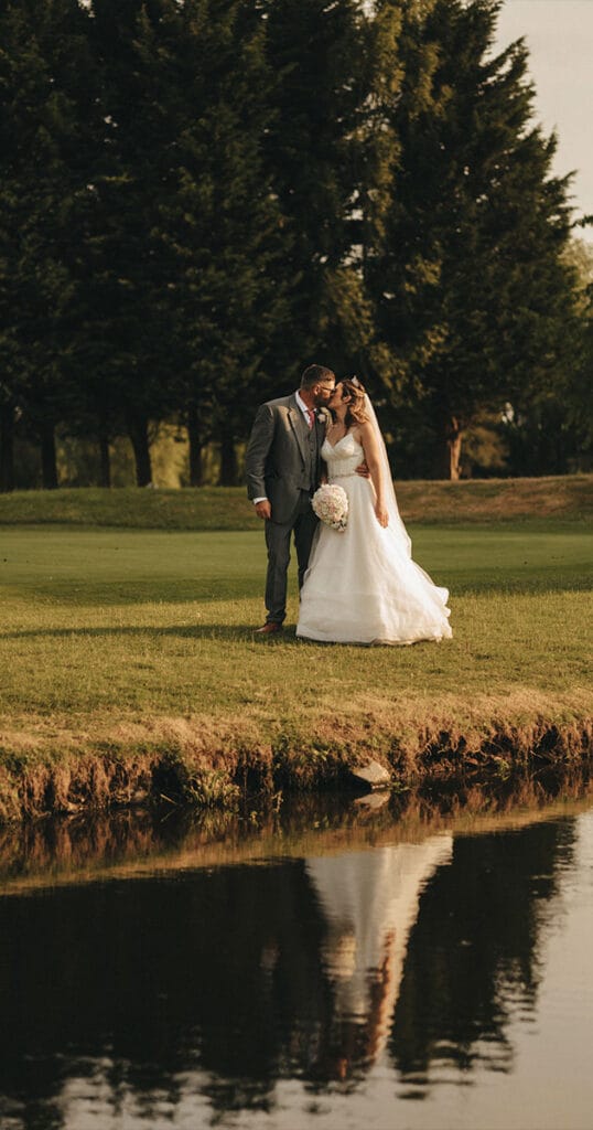 A bride and groom share a kiss on a grassy area at Messingham Grange near a tranquil pond. The bride is in a white dress holding a bouquet, while the groom dons a dark suit. Tall trees create a lush backdrop, their reflection beautifully mirrored in the calm water. © Aimee Lince Photography