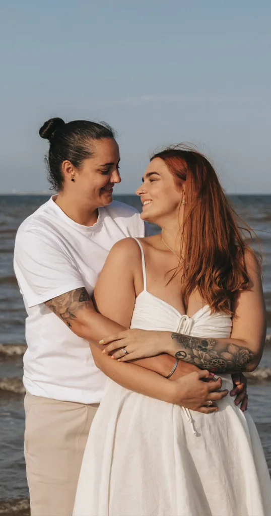 A couple embraces on Cleethorpes Beach. The person on the left has long hair tied back, wearing a white t-shirt adorned with arm tattoos. Their partner, with long red hair and a white dress, smiles warmly. The tranquil sea and sky compose the picturesque backdrop. © Aimee Lince Photography - Wedding photographer in Lincolnshire, Yorkshire & Nottinghamshire