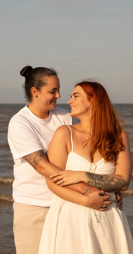 On the serene Cleethorpes beach, two people stand embracing and smiling at each other. One sports a white t-shirt, tattoos, and a bun; the other flaunts long red hair, tattoos, and a white dress. With the ocean and clear sky as their backdrop, it's an idyllic scene perfect for engagement photography. © Aimee Lince Photography - Wedding photographer in Lincolnshire, Yorkshire & Nottinghamshire