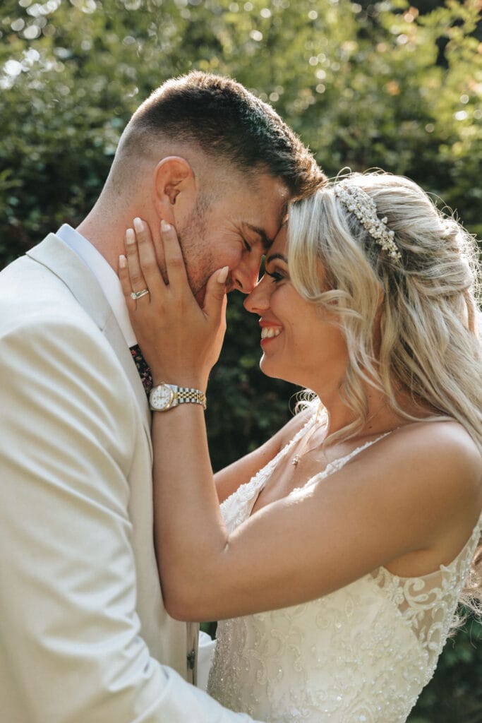 A couple shares an intimate moment outdoors at the scenic Mount Pleasant Hotel. The woman, in a white lace wedding dress and jeweled headpiece, gently holds the man's face. He is dressed in a light-colored suit with a watch. Both smile with their foreheads touching, surrounded by lush greenery. © Aimee Lince Photography