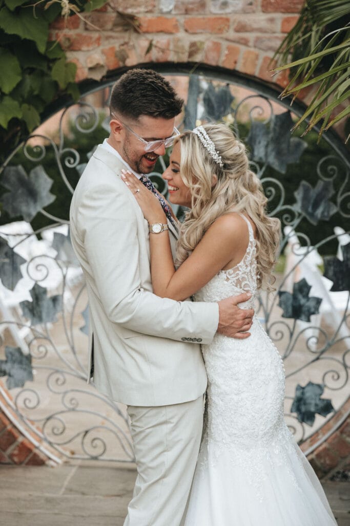 A couple embraces on their wedding day at the enchanting Mount Pleasant Hotel. The groom, in a light beige suit and glasses, smiles lovingly at his bride in a lace-embroidered gown and tiara. They stand blissfully before a decorative wrought iron gate and brick wall adorned with greenery. © Aimee Lince Photography