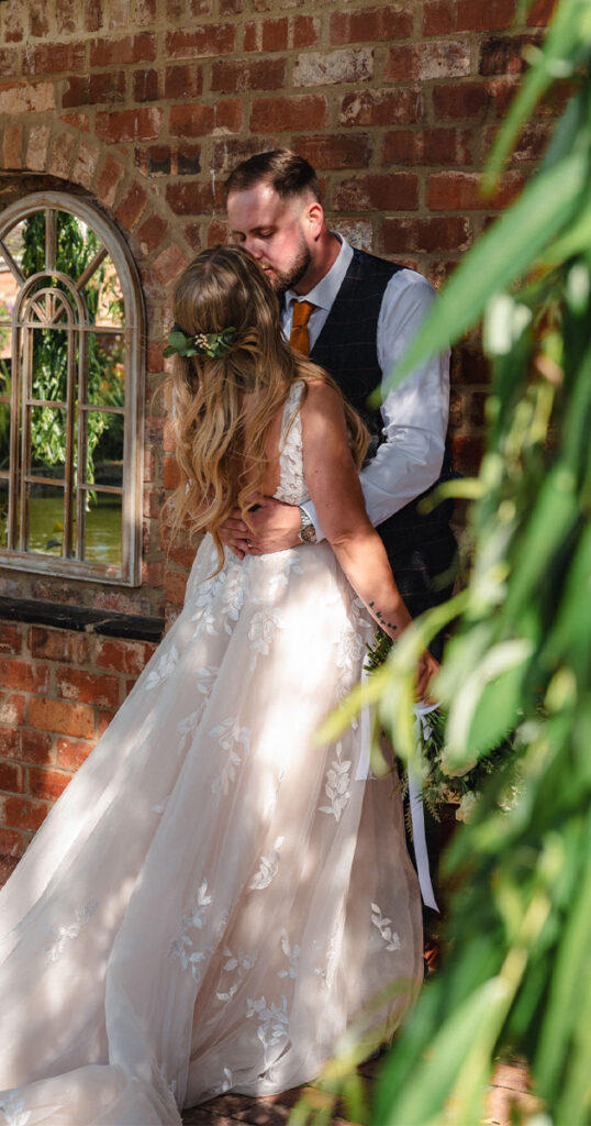 A newlywed couple shares a kiss by a brick wall with arched windows in Stallingborough. The bride wears a flowing white gown with floral patterns and a flower crown, while the groom dons a gray vest, white shirt, and orange tie. Green foliage frames the intimate, sunlit scene. © Aimee Lince Photography