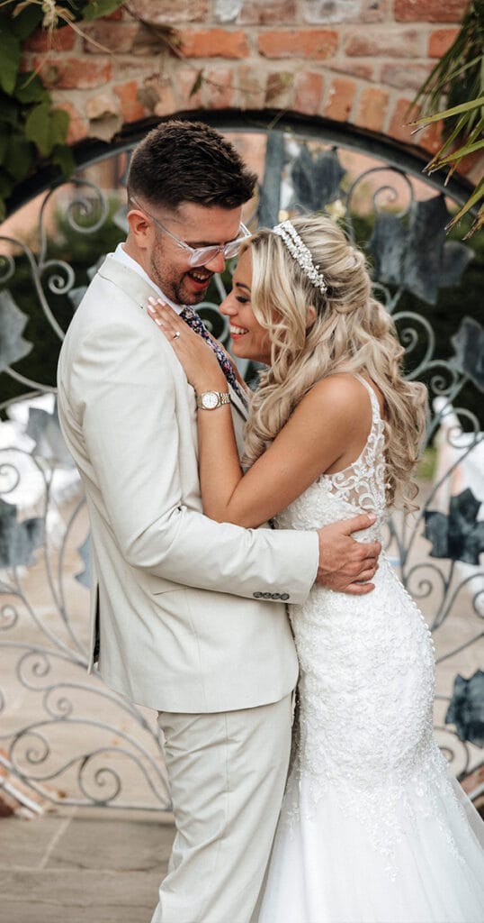 A bride and groom share a joyful embrace at the entrance of the ornate, wrought-iron gate of Mount Pleasant Hotel. The bride wears a white lace gown and headpiece, while the groom is in a light beige suit. Both are smiling warmly, creating an atmosphere of happiness and celebration amid lush greenery. © Aimee Lince Photography