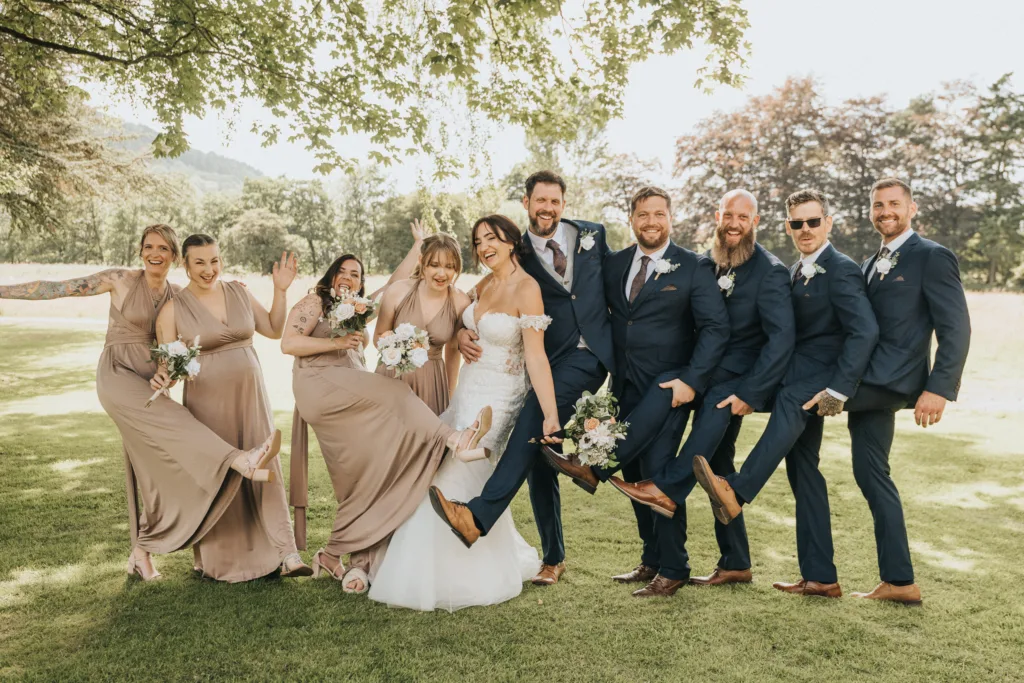 A joyful wedding party poses outside on grass, laughing. Like fun family portraits, bridesmaids in taupe and groomsmen in navy kick up one leg alongside the bride and groom, holding bouquets under leafy trees in bright daylight. © Aimee Lince Photography - Wedding photographer in Lincolnshire, Yorkshire & Nottinghamshire