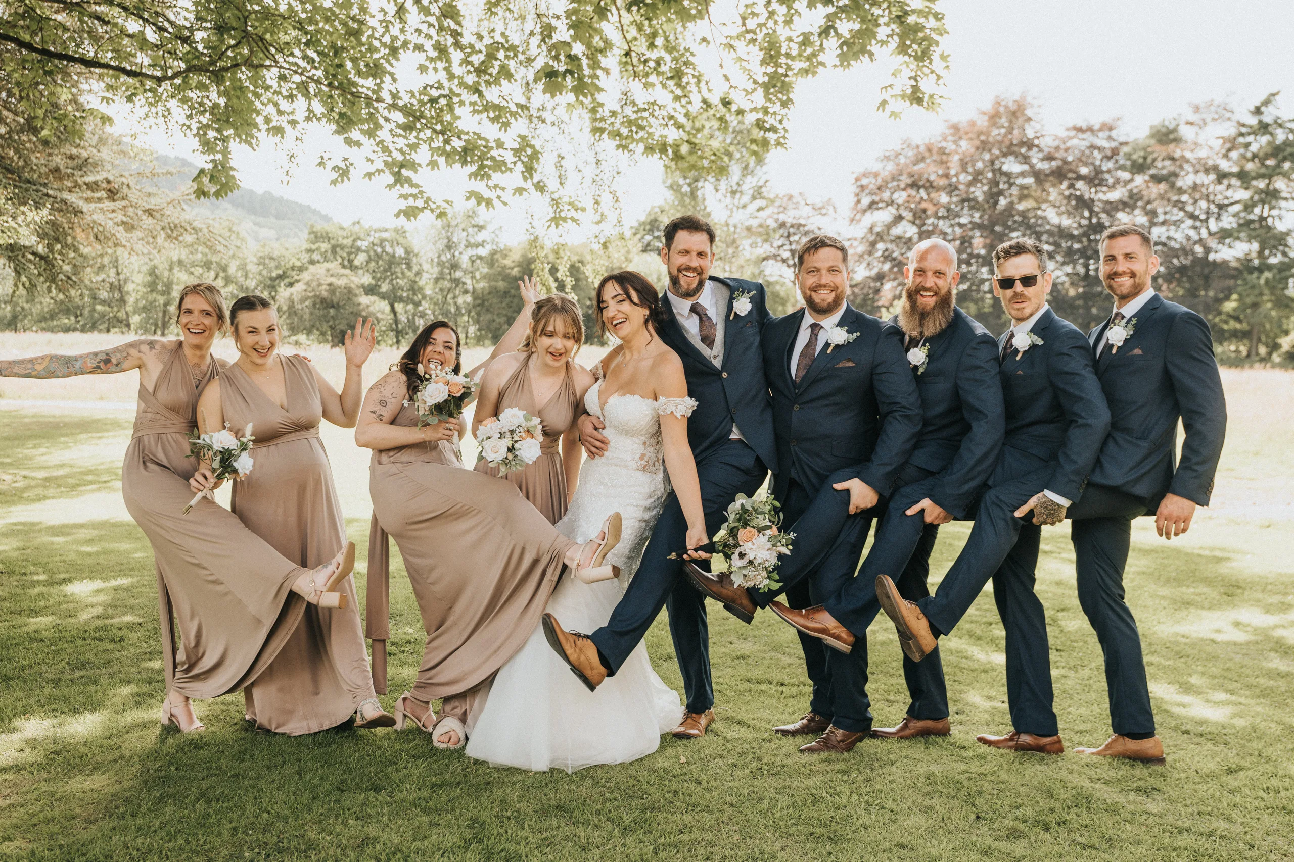 carina-shane-wedding-photo-scarborough-1-of-1-©-aimee-lince-photography-16-07-2025 A joyful wedding party poses outside on grass, laughing. Like fun family portraits, bridesmaids in taupe and groomsmen in navy kick up one leg alongside the bride and groom, holding bouquets under leafy trees in bright daylight. © Aimee Lince Photography - Wedding photographer in Lincolnshire, Yorkshire & Nottinghamshire