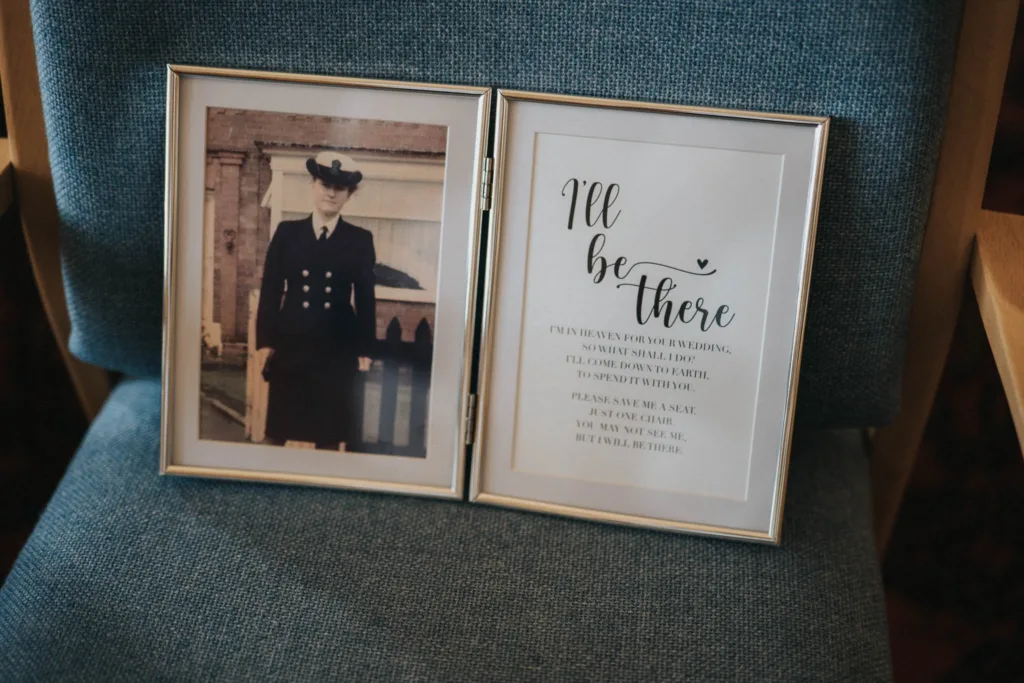 Absent Friends - Kirsty & John A framed photograph on a chair features a person in a naval uniform alongside the poem "I'll be there," honoring absent friends by expressing their presence in spirit at weddings and special events. The metallic frames rest against a textured blue fabric backdrop. © Aimee Lince Photography - Wedding photographer in Lincolnshire, Yorkshire & Nottinghamshire