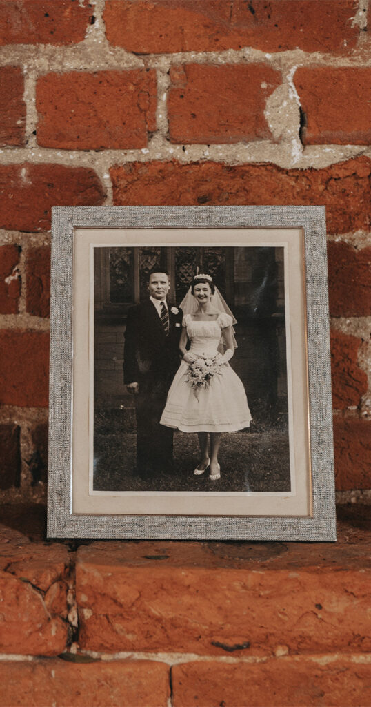 A black-and-white photo of a bride and groom is framed and displayed on a red brick shelf. The bride, honoring absent friends with her bouquet, wears a short wedding dress beside the suited groom. The silver frame boasts a textured border against the backdrop of grass and brick wall. © Aimee Lince Photography