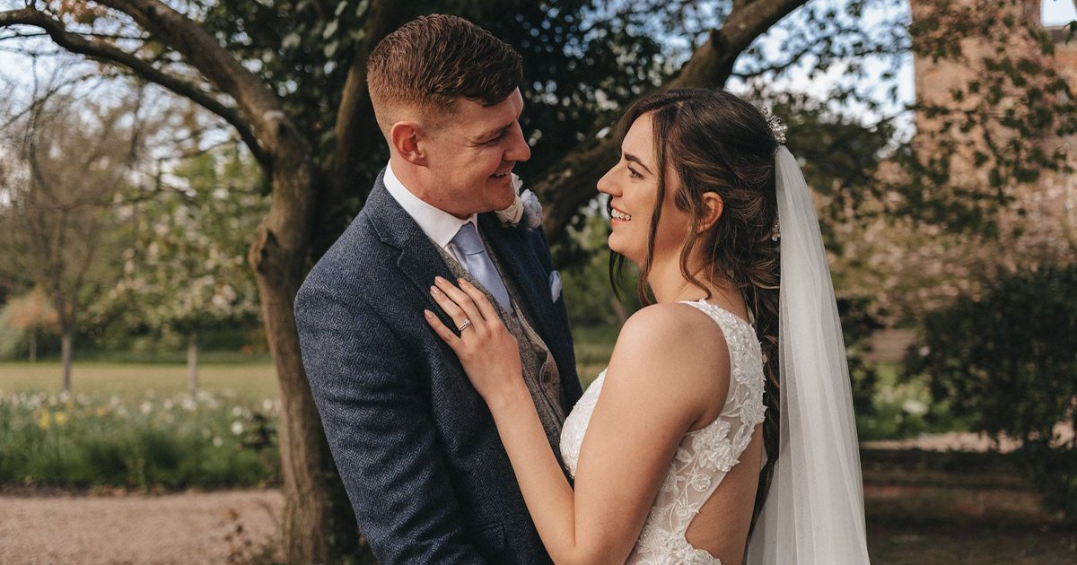 A bride and groom stand outdoors at their Hodsock Priory wedding, smiling at each other. The bride wears a white lace dress and veil, whilst the groom wears a navy suit. Trees and greenery provide a stunning spring wedding backdrop. © Aimee Lince Photography, wedding and lifestyle photographer in Lincolnshire