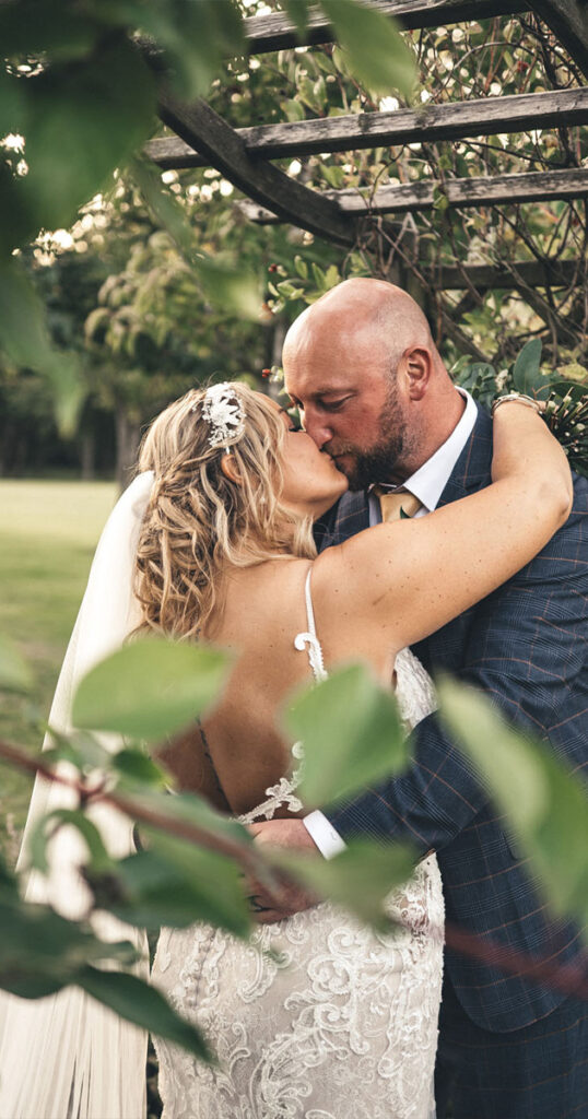 A couple kisses under a garden archway at Laceby Manor. The bride, in a lace wedding dress and veil, wears a decorative hairpiece. The groom, in a checked suit and yellow tie, holds her closely. Leaves and greenery frame the foreground, adding a natural, romantic setting to the scene. © Aimee Lince Photography