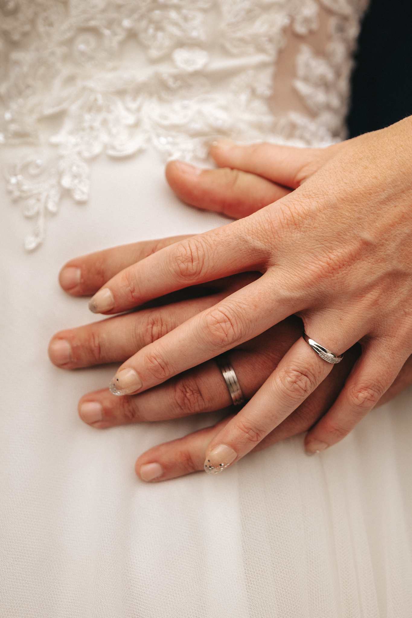 Close-up of two hands with wedding rings overlaid on a white lace bridal dress at The Admiral Rodney Hotel. The top hand, with a delicate ring featuring a stone, rests on another hand, which wears a simple, smooth band. Both hands have manicured nails, and the intricate lace detailing of the dress is visible in the background. © Aimee Lince Photography