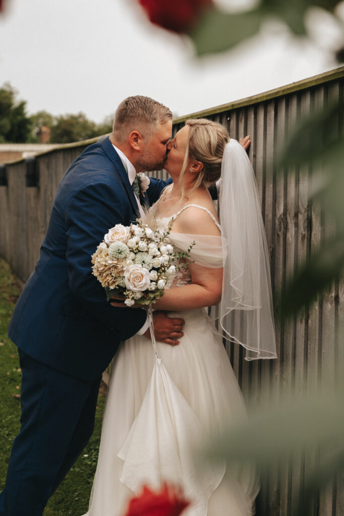 A newlywed couple shares a romantic kiss by a wooden fence at The Admiral Rodney Hotel. The groom is dressed in a blue suit, while the bride wears an off-the-shoulder white wedding dress and veil, holding a bouquet of white and pastel flowers. Blurred red flowers in the foreground add depth to the image. © Aimee Lince Photography