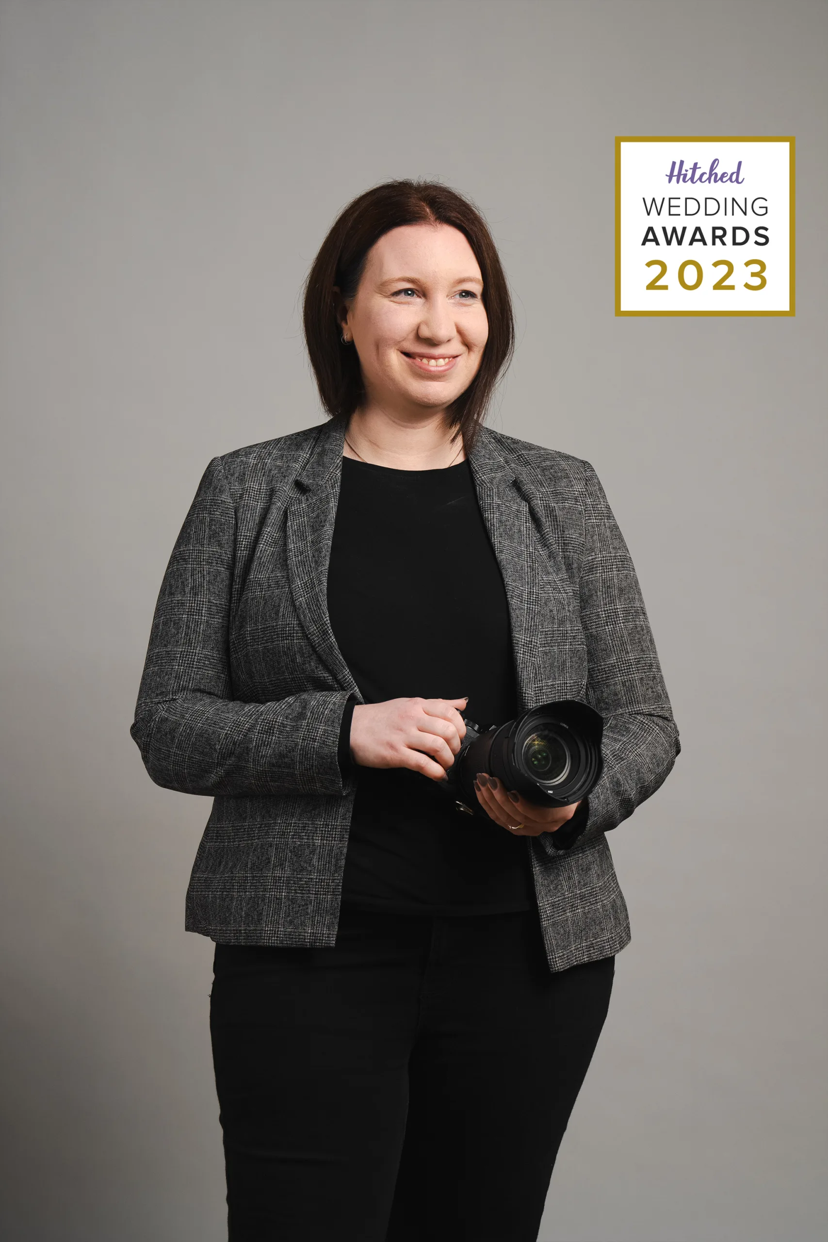 A wedding photographer holding a camera, dressed in a business casual outfit, beams with pride at the 2023 hitched wedding awards, showcasing her award recognition in the industry.