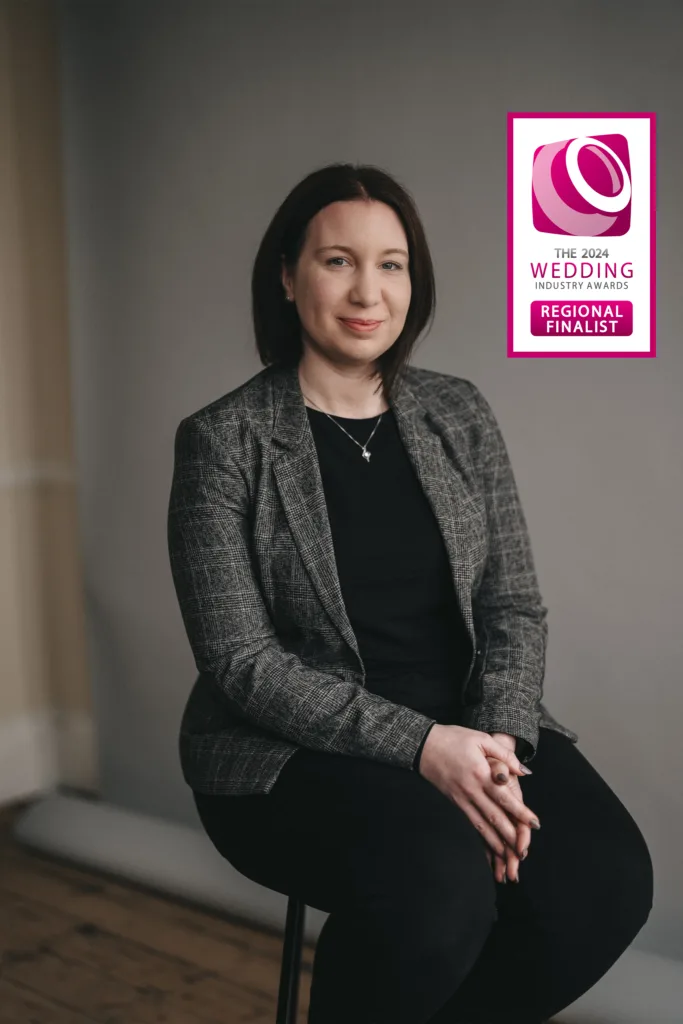 A poised wedding photographer in a herringbone blazer and black attire sits comfortably on a stool, radiating confidence and professionalism, with a badge indicating she is a "2024 wedding industry regional finalist