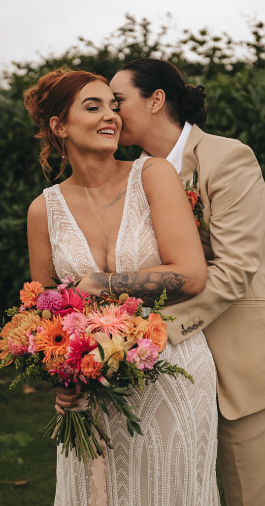 At a wedding, a bride in a white gown holds a vibrant bouquet of pink, orange, and yellow flowers. Another person in a tan suit with a boutonniere lovingly whispers in her ear. They stand outdoors, surrounded by lush greenery, sharing a joyous and intimate moment together. © Aimee Lince Photography