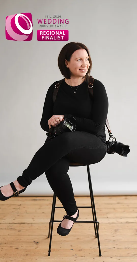 A woman with shoulder-length brown hair sits on a tall stool against a plain gray background. She wears a black outfit and holds a camera, with another across her shoulder. A badge in the top-left corner reads, "Wedding Photographer of the Year 2023 Regional Finalist." The floor is wooden. © Aimee Lince Photography - Wedding photographer in Lincolnshire, Yorkshire & Nottinghamshire